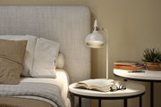 White metal floor lamp beside bed with beige-grey cushions and bedside table with books and plant