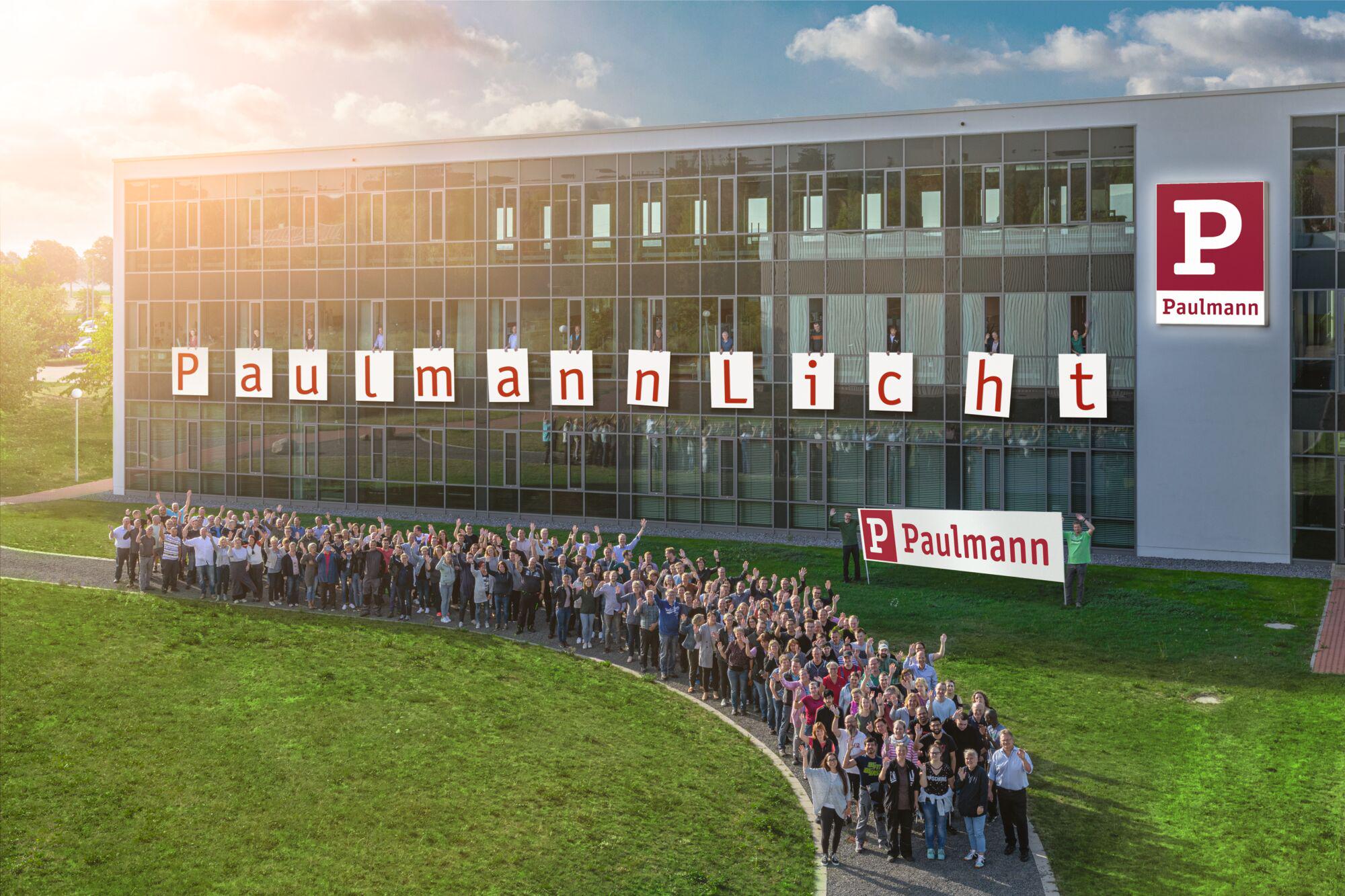 Große Gruppe Mitarbeiter vor Paulmann Firmengebäude mit Schriftzug Paulmann Licht und Firmenlogo.