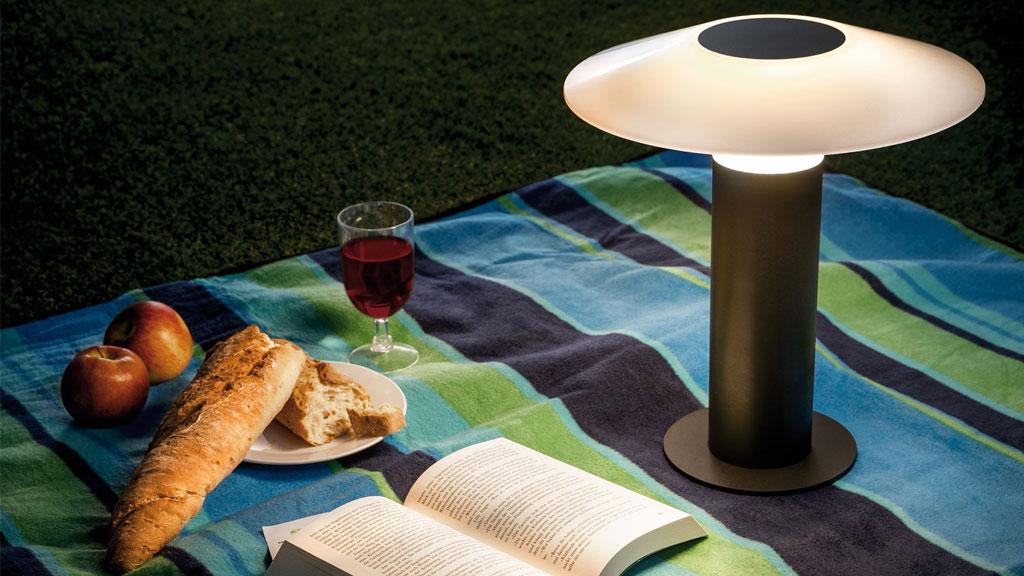 Moderne Tischlampe mit schwarzem Metallfuß und weißem Lampenschirm auf Picknickdecke mit Brot und Wein.