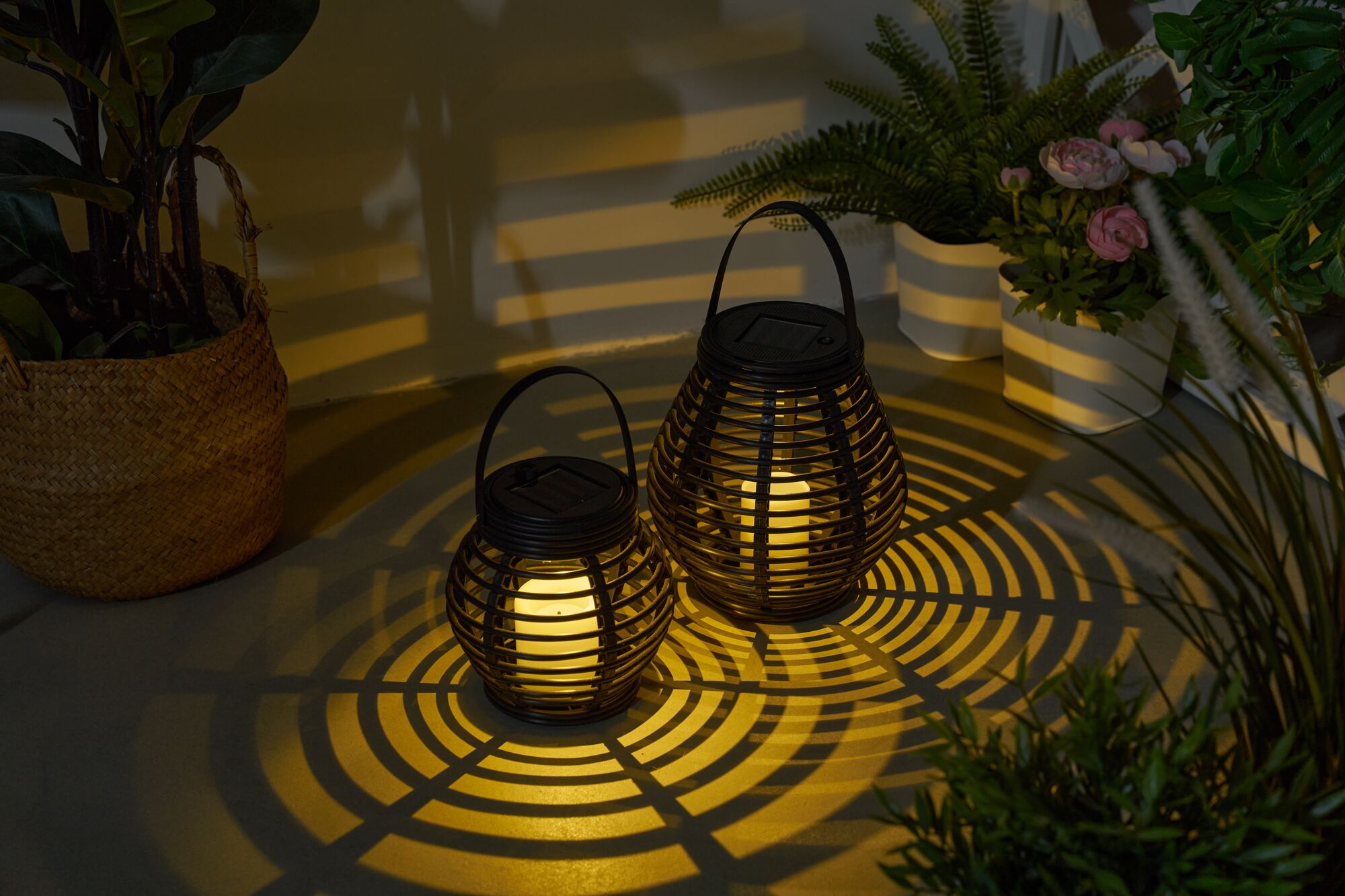 Two black metal solar lanterns with warm white LED light casting decorative shadows in a garden setting.
