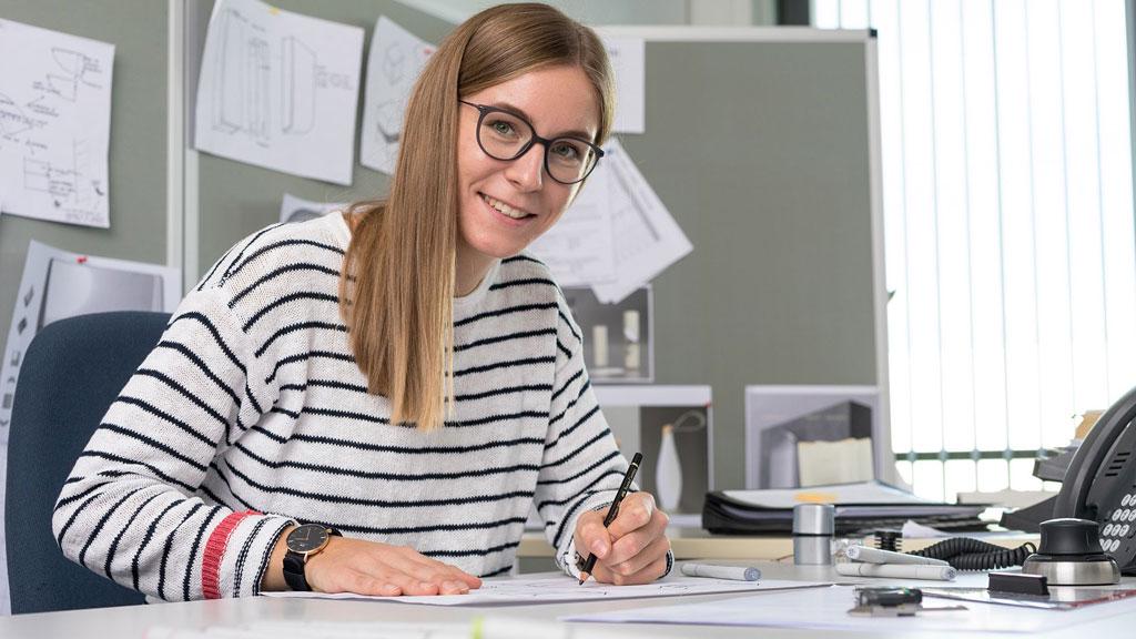 Junge Frau mit Brille und gestreiftem Pullover arbeitet an technischen Zeichnungen im Büro.