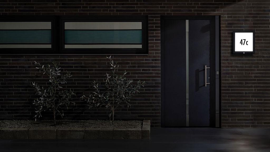 Moderne indgang med mørk dør og oplyst husnummer på murstensvæg om natten