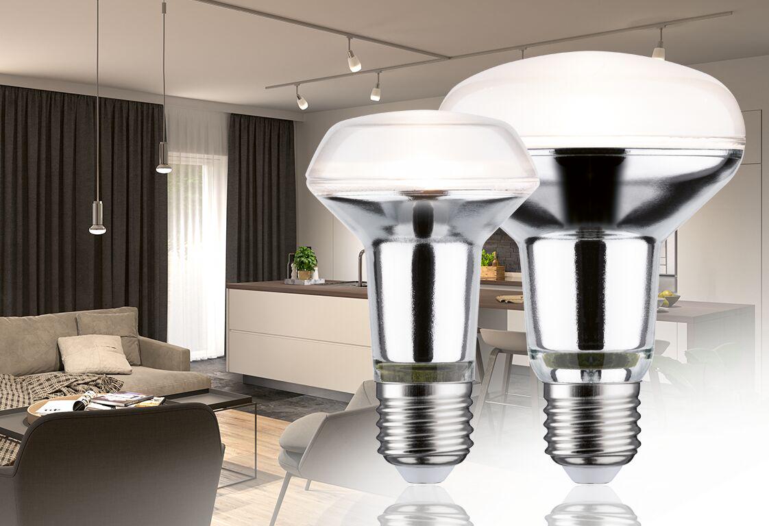 Zwei reflektierende Glühlampen mit E27-Fassung in Weiß und Silber vor modernem Wohnzimmer mit Beleuchtung.