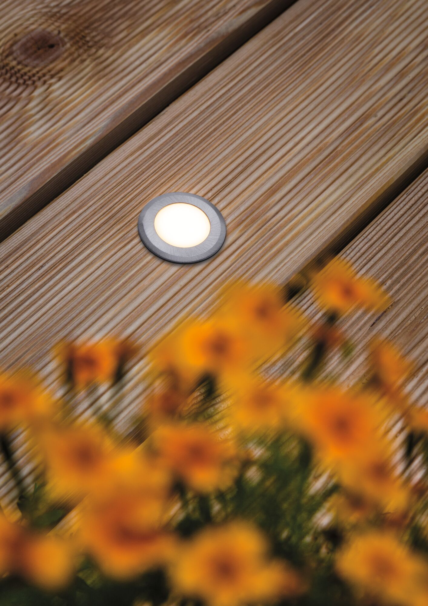 Round LED recessed floor light in brushed aluminum on wooden terrace with flowers in foreground