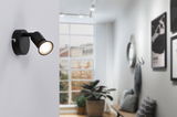 Black metal LED wall spotlight with adjustable head in modern living space with large window.