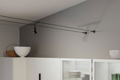 Black cable track ceiling spotlight in modern room above white kitchen cabinet with dishes.