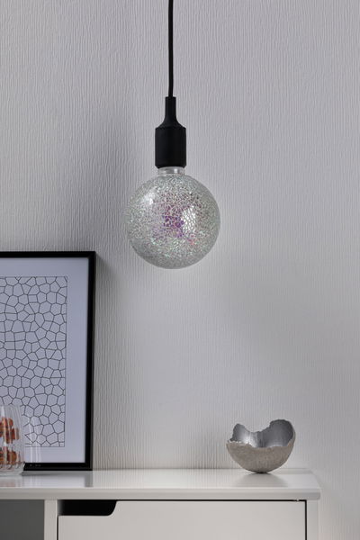 Hangende gloeilamp met zilveren mozaïekglas en zwarte kabel tegen witte muur.