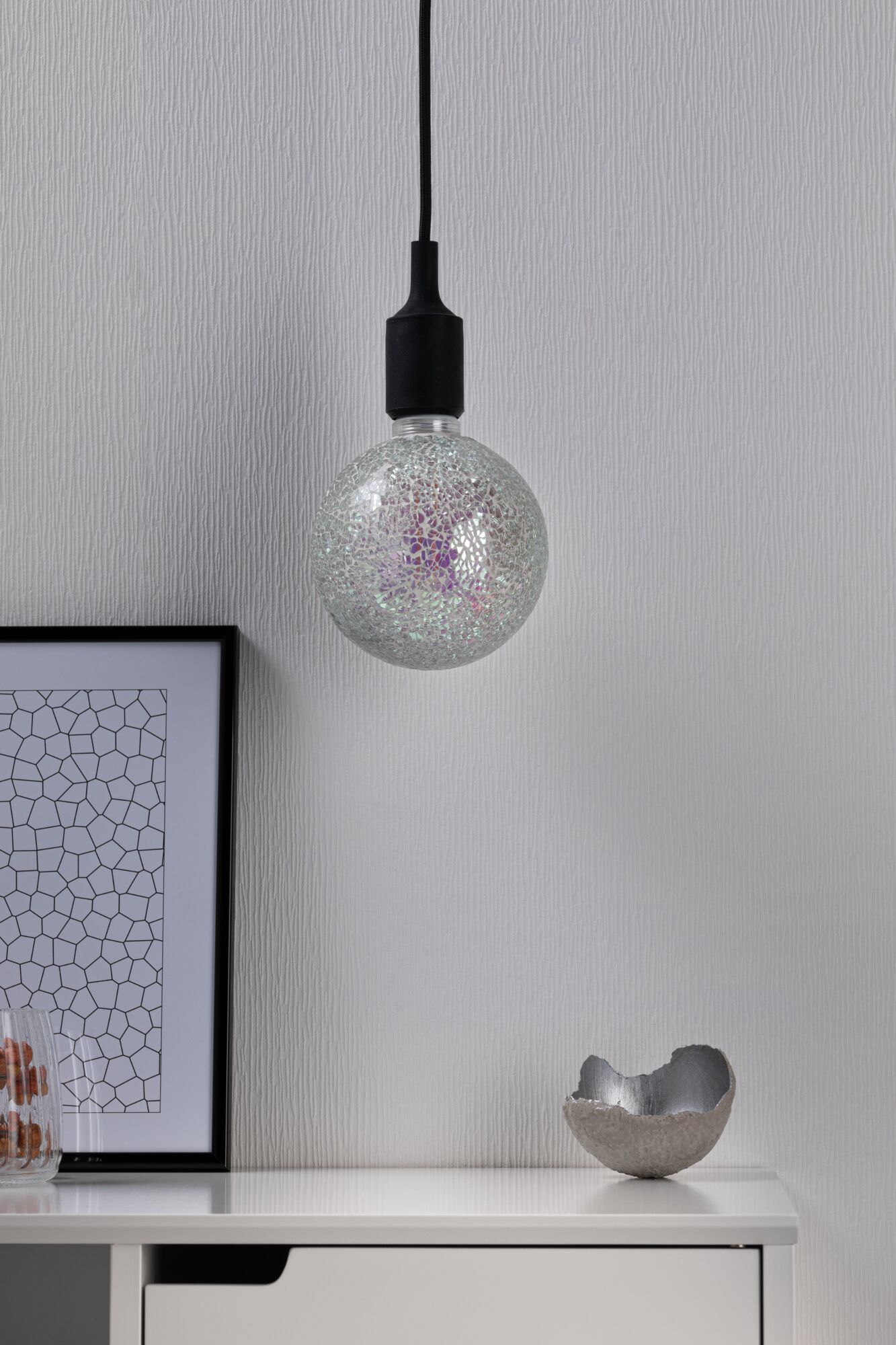 Hangende gloeilamp met zilveren mozaïekglas en zwarte kabel tegen witte muur.