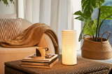 Cylindrical white patterned table lamp on wicker table near sofa and plant in cozy living room.