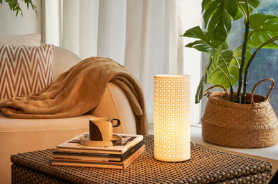 Cilindervormige witte tafellamp met patroon op gevlochten tafel naast bank en plant in woonkamer.