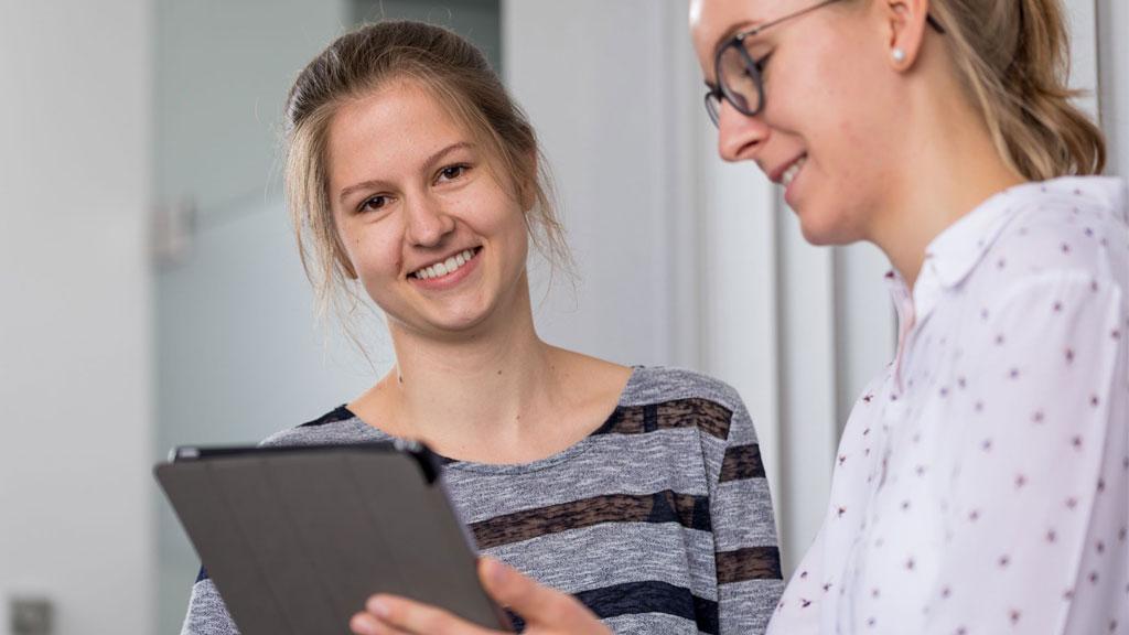 Zwei junge Frauen im Gespräch mit Tablet, freundliche Arbeitsatmosphäre im Büro, Karriere und Ausbildung.