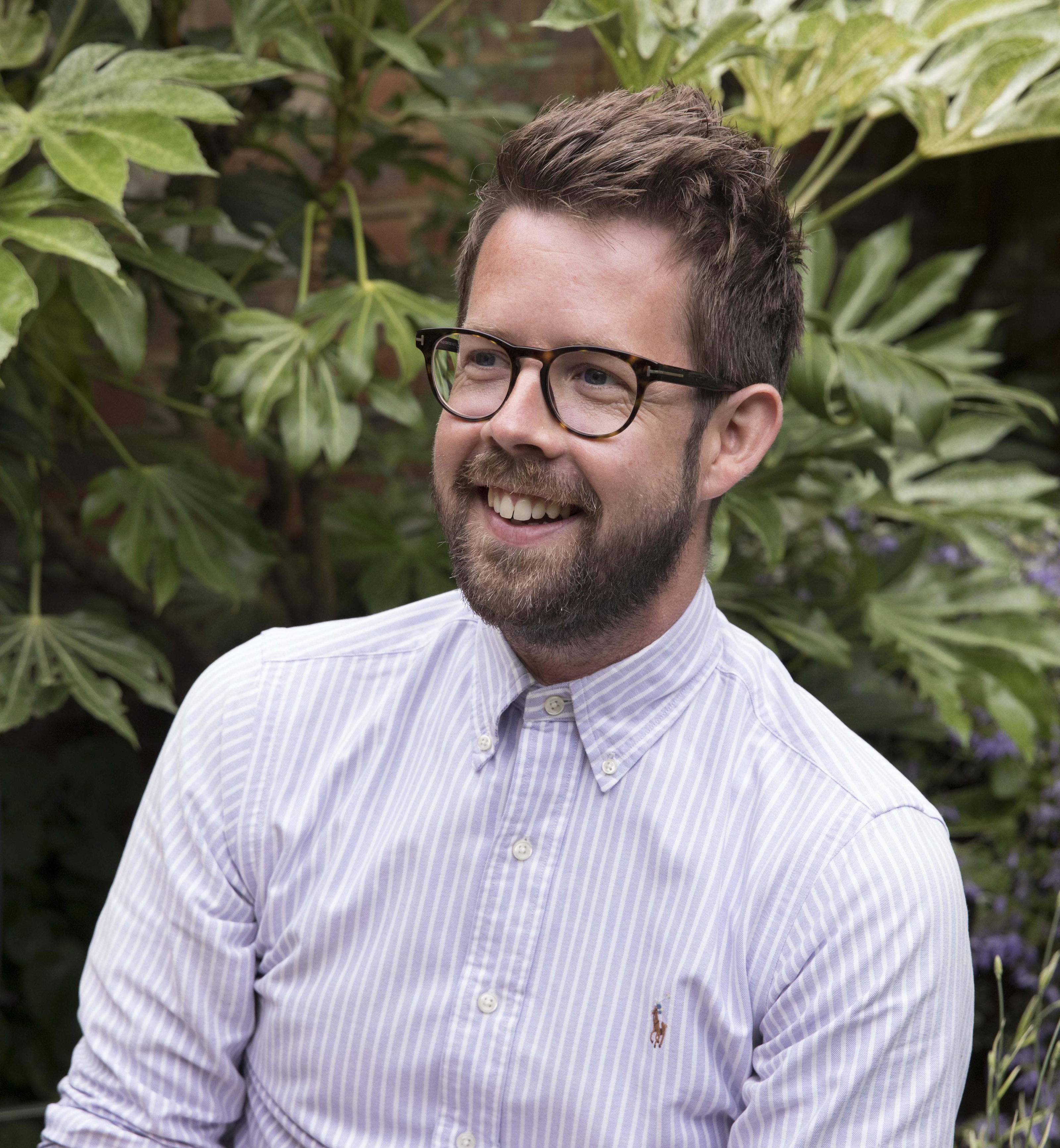 Portrait of a smiling man with glasses and striped shirt in front of green foliage background