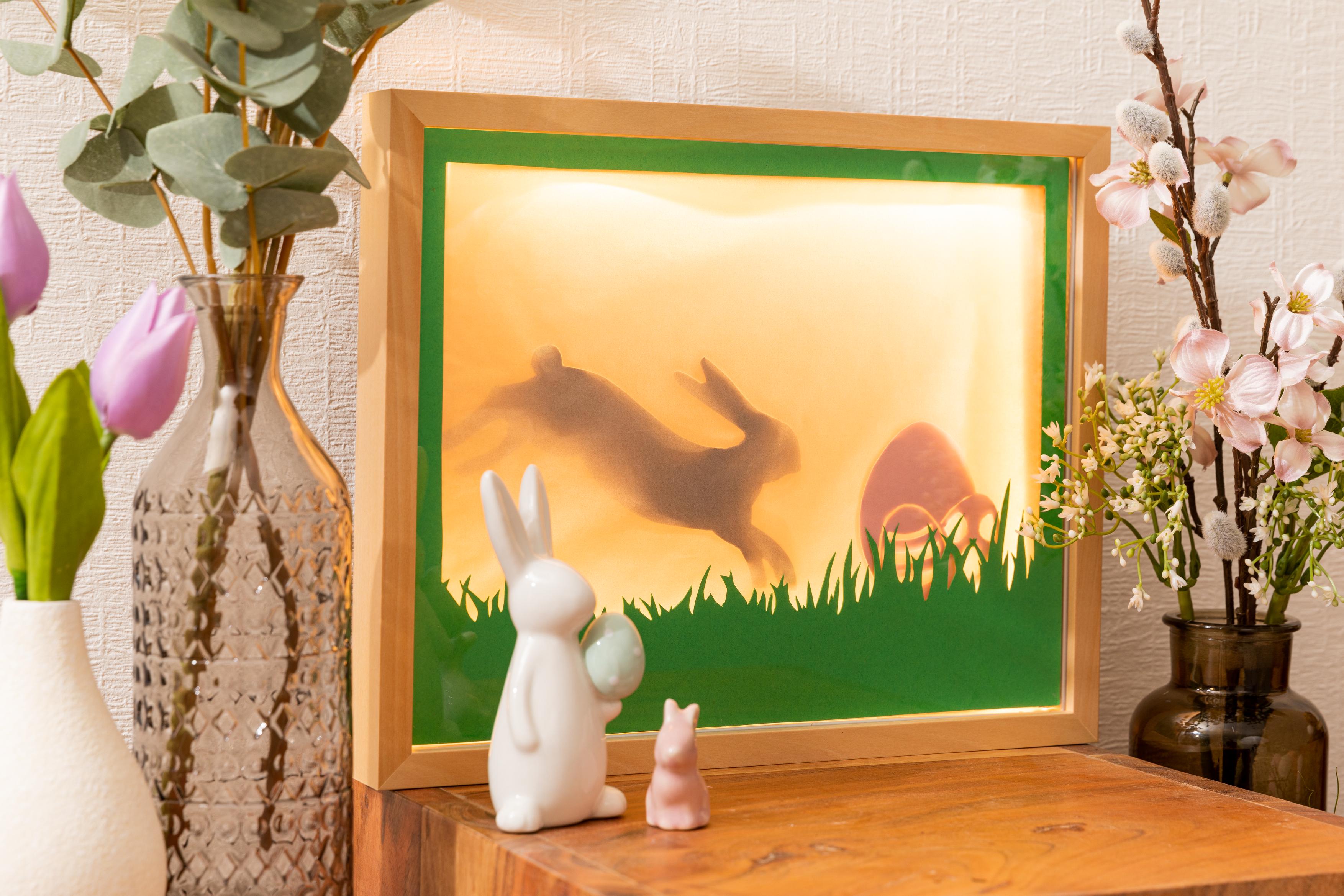 Osterdekoration mit beleuchtetem Holzrahmen, Hasenschatten und Keramikhasen auf Holztisch mit Blumen.