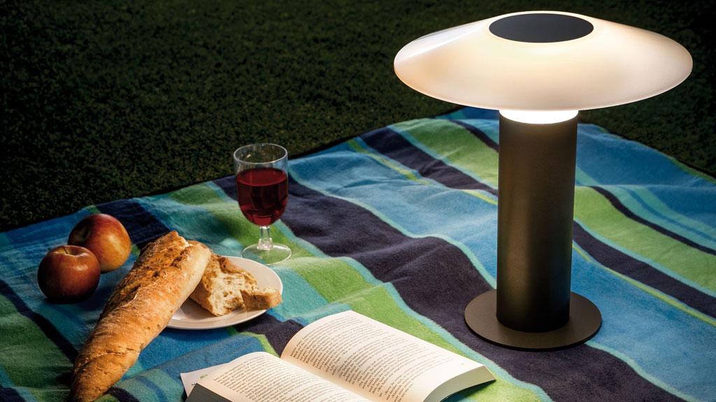 Modern black garden lamp with white shade on picnic blanket with bread, apples, and book.