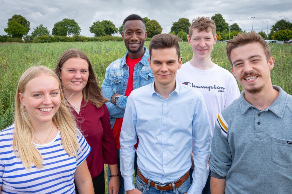 Six young adults smiling outdoors in front of green field, representing team spirit and training.