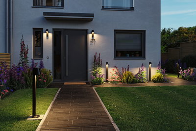 Modern black outdoor wall and bollard lights illuminate the house entrance with garden path and flower beds.