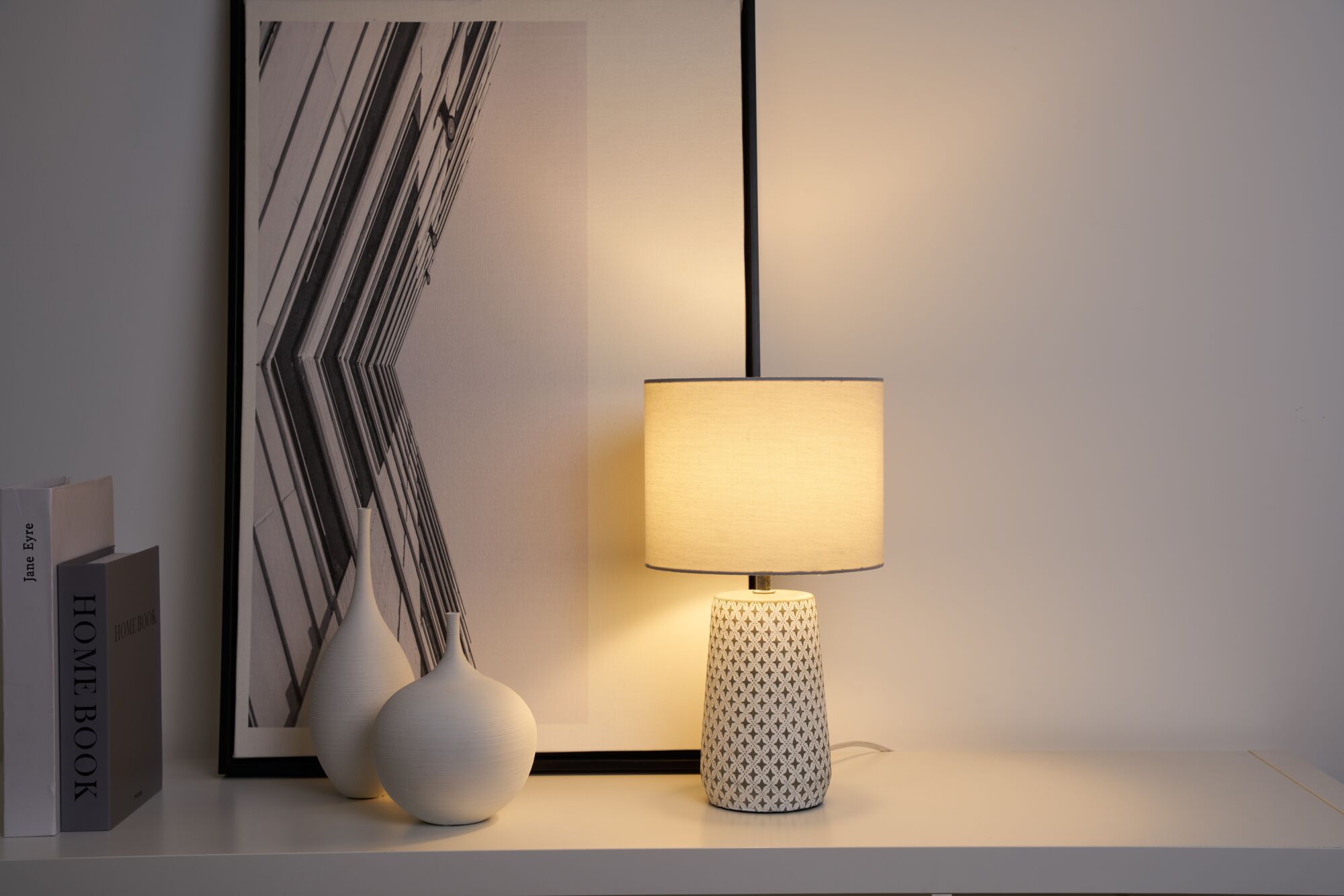 Table lamp with patterned ceramic base and light fabric shade on white shelf with decor and artwork.