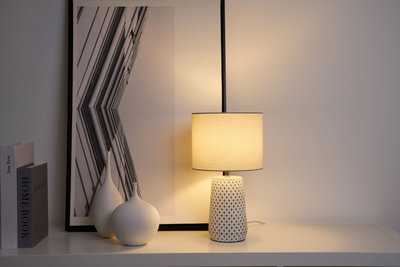Lampe de table avec base en céramique à motif et abat-jour en tissu clair sur étagère blanche avec déco.