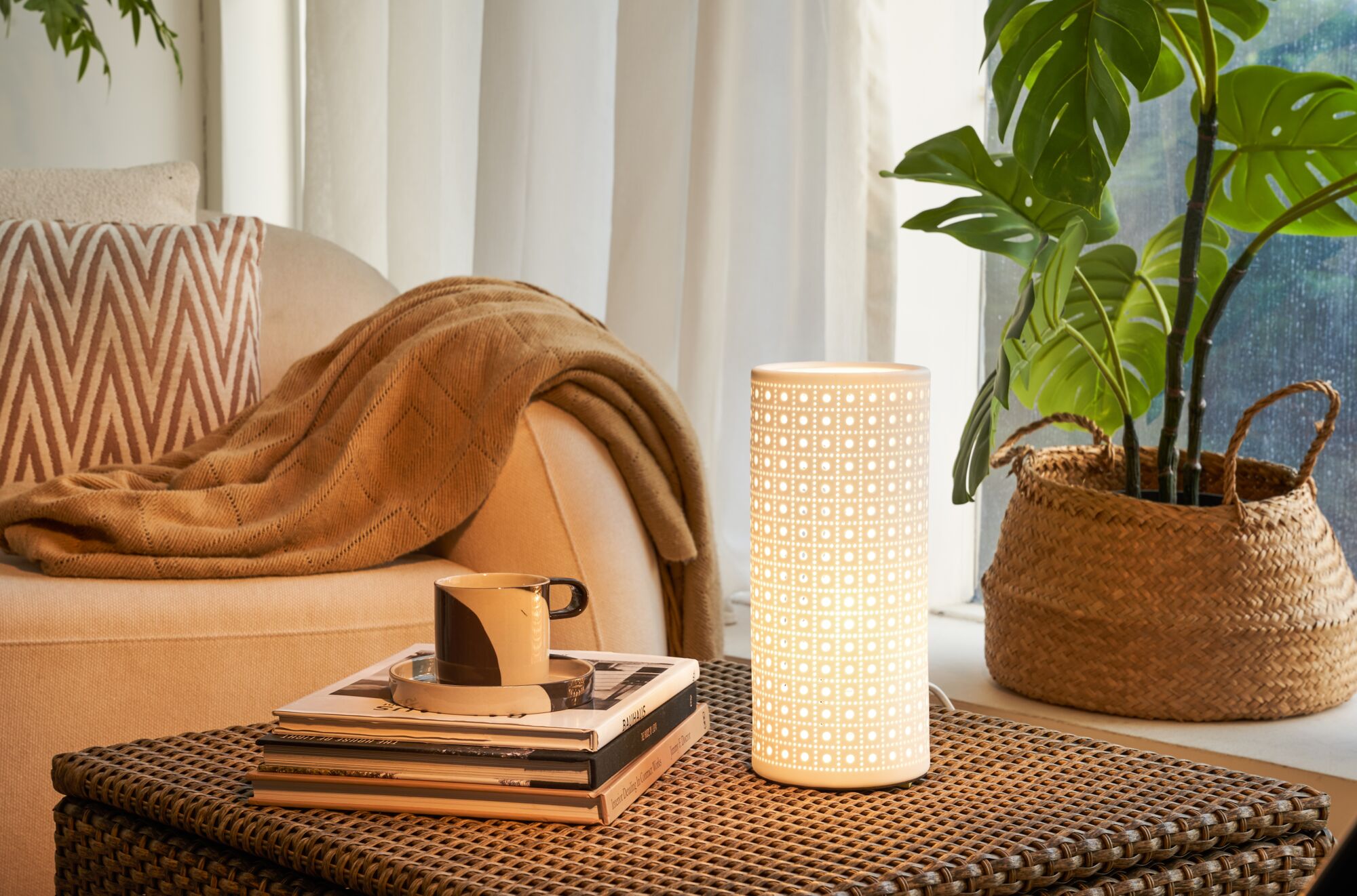 White ceramic table lamp with geometric pattern on woven table near sofa and plant in cozy room