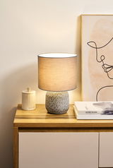 Tischlampe mit grauem Stoffschirm und gemustertem Keramikfuß auf Holzkommode neben Büchern und Deko.