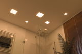 Moderne vierkante LED-plafondlampen in warm wit verlichten een douche met glazen wand en houten jaloezieën.