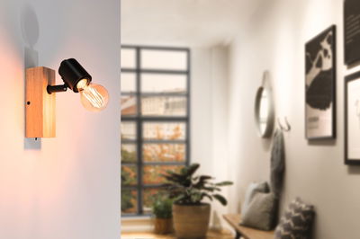 Wall lamp with wooden base and black metal head, warm light in modern living space with window view.