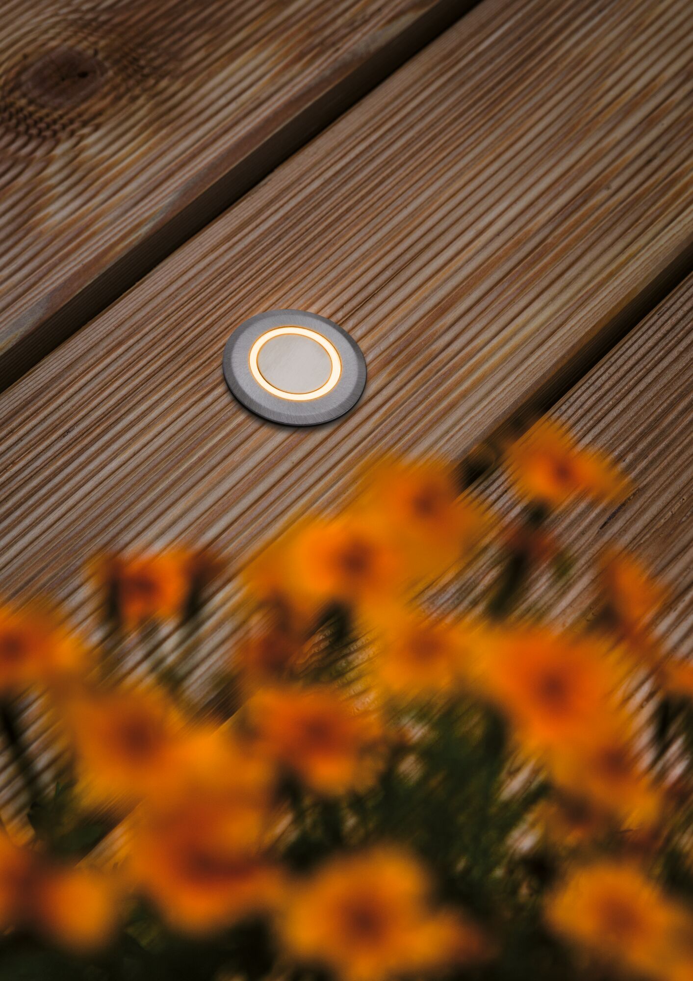 Round LED recessed light in brushed aluminum with warm white glow on wooden terrace.