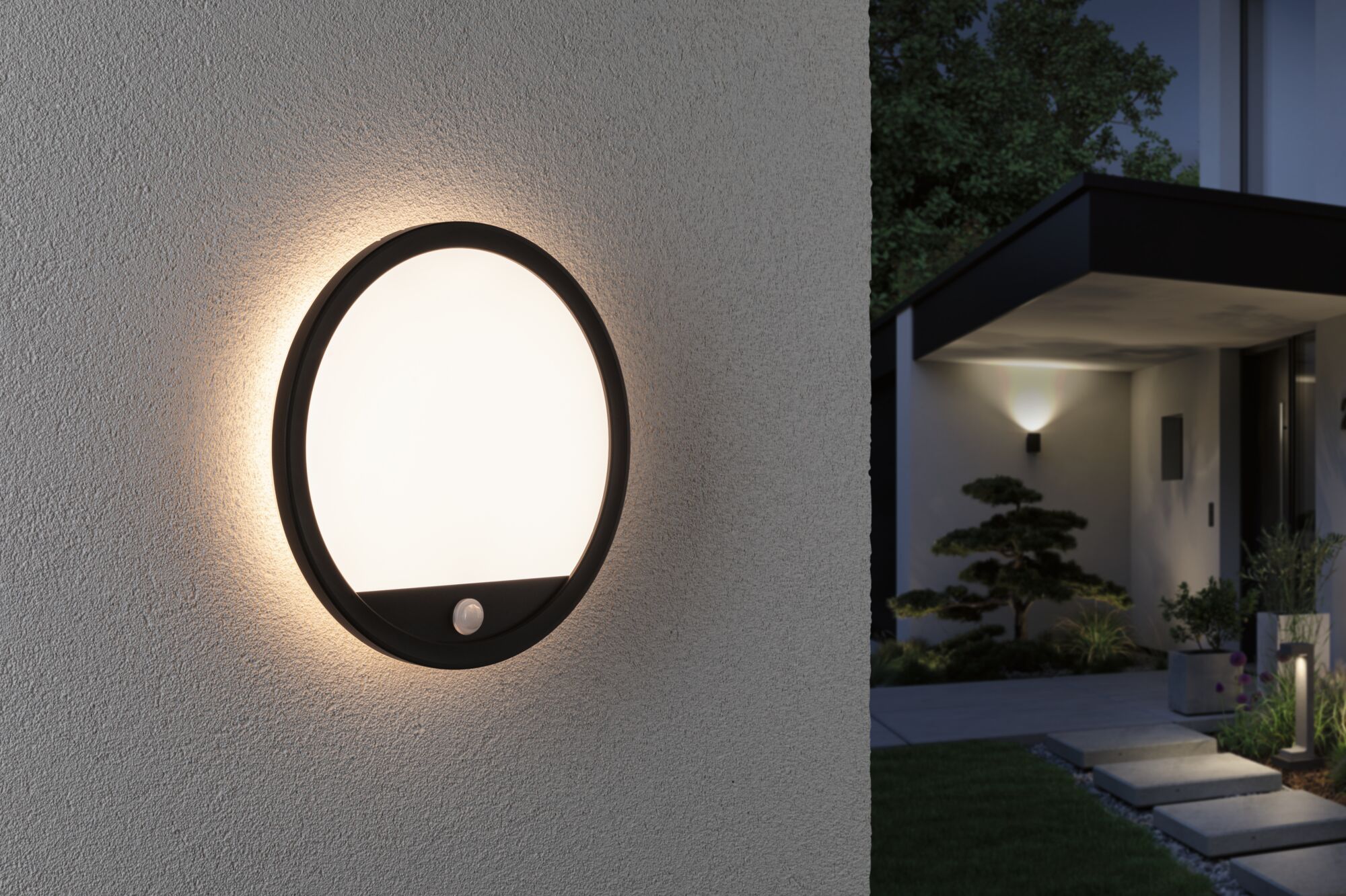 Round outdoor wall light with black frame and warm white glow on house exterior at night