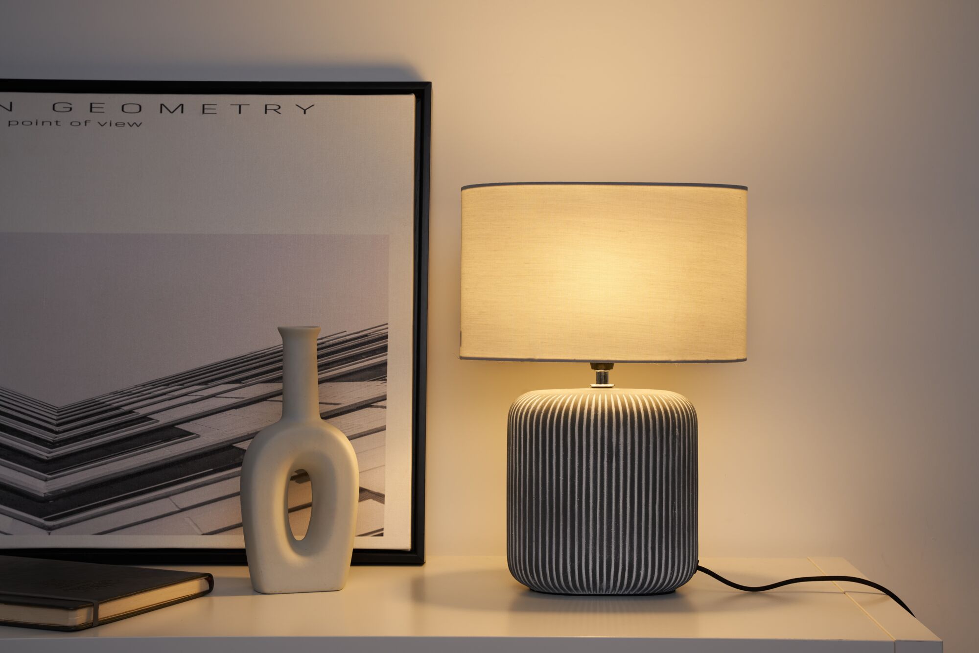 Modern table lamp with grey ceramic base and light fabric shade on white shelf with decor.