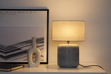 Modern table lamp with grey ceramic base and light fabric shade on white shelf with decor.