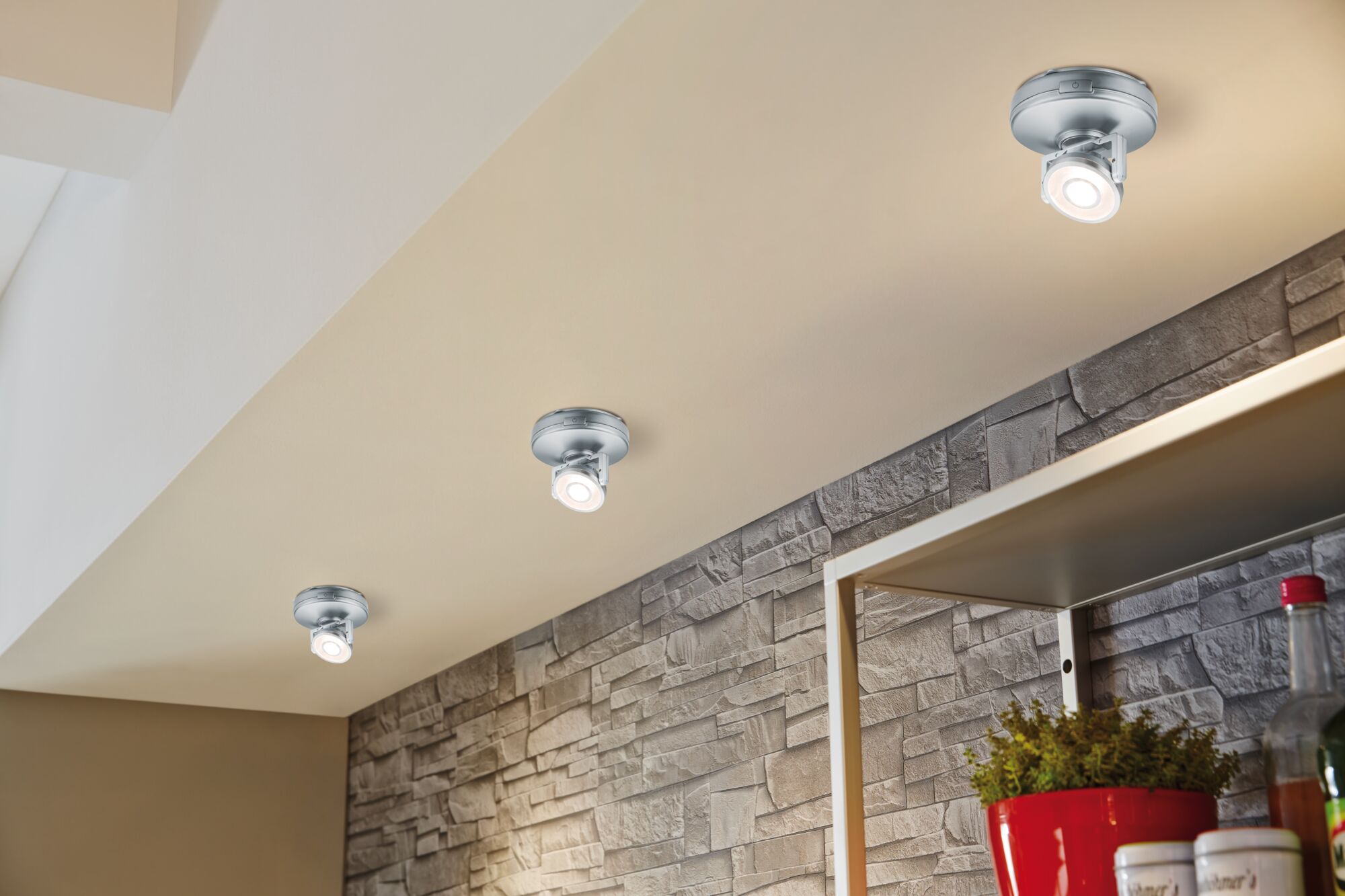 Trois spots LED argentés au plafond beige avec mur en pierre grise et pot de fleurs rouge.