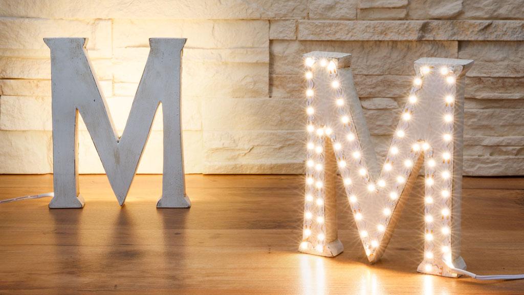 Two white metal letter M signs, one with warm white LED lights, on wooden floor against stone wall.