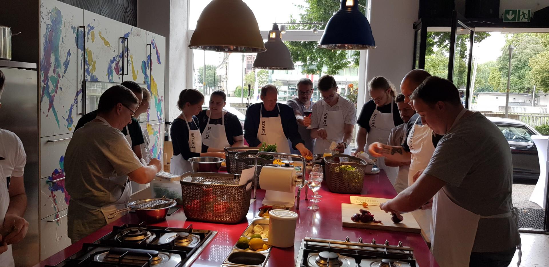 Gruppe von Menschen mit weißen Janus-Schürzen beim gemeinsamen Kochen in moderner Küche mit bunten Schränken.
