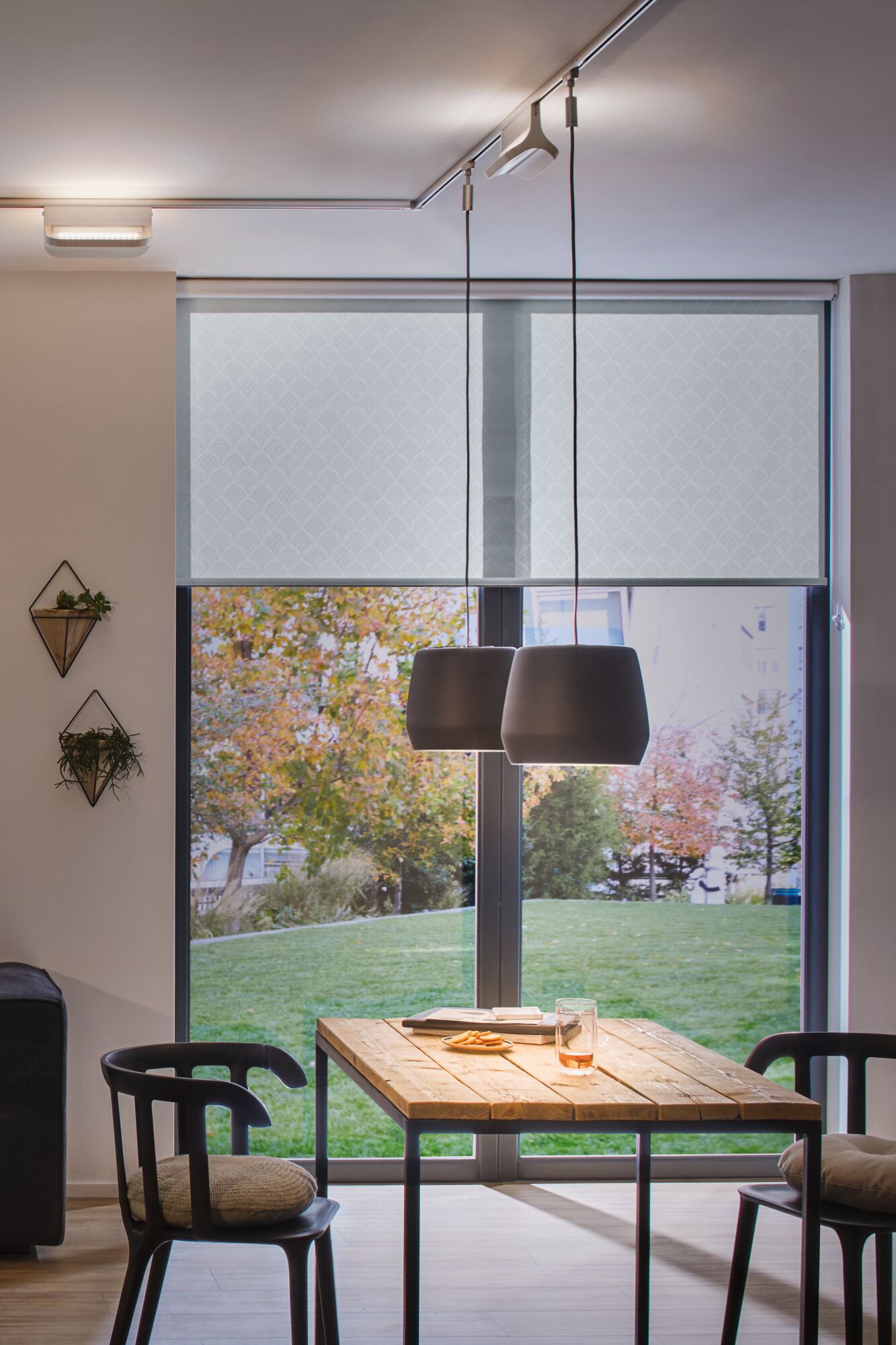 Modern dining area with wooden table, black chairs, and two gray pendant lamps in front of large windows with patterned blinds