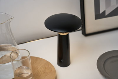 Black metal LED table lamp with round shade on white table near glass and wooden tray