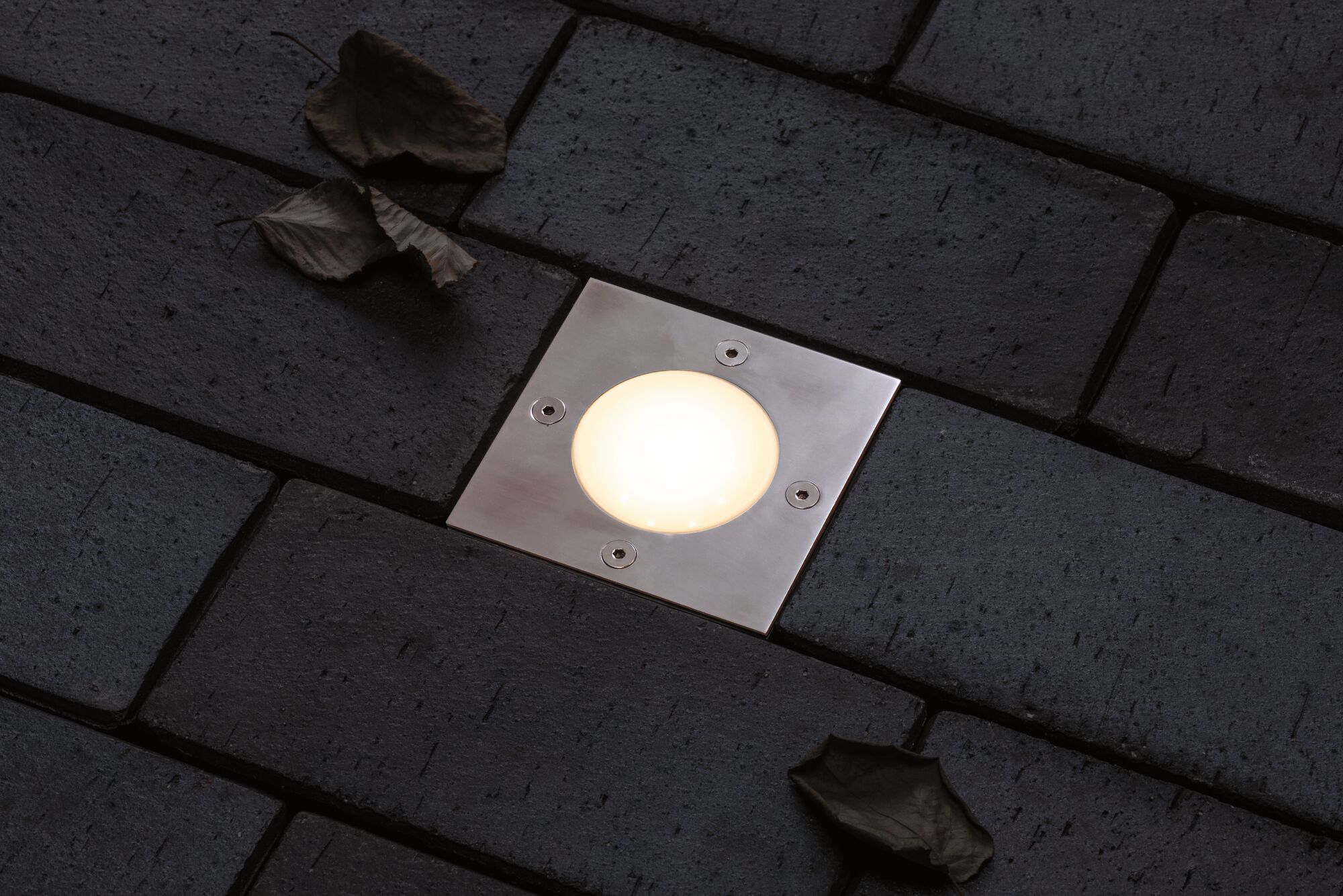 Recessed square LED floor light in stainless steel with warm white glow on dark pavement surface.