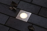 Recessed square LED floor light in stainless steel with warm white glow on dark pavement surface.