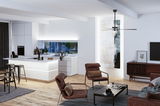 Salon moderne avec fauteuils en tissu marron, sol en bois et cuisine blanche ouverte avec éclairage LED.