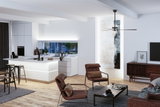 Modernes Wohnzimmer mit braunen Stoffsesseln, Holzfußboden und offener weißer Küche mit LED-Beleuchtung.