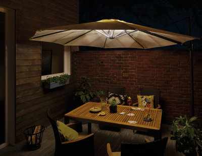 Cream-colored patio umbrella with LED lights over wooden table and chairs on terrace at night