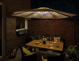 Cream-colored patio umbrella with LED lights over wooden table and chairs on terrace at night