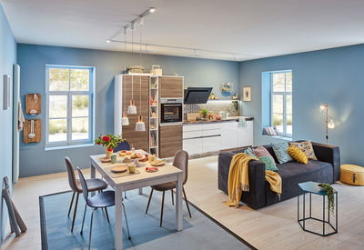 Modern open living room with blue walls, white dining table, gray sofa, and wood kitchen with LED lighting