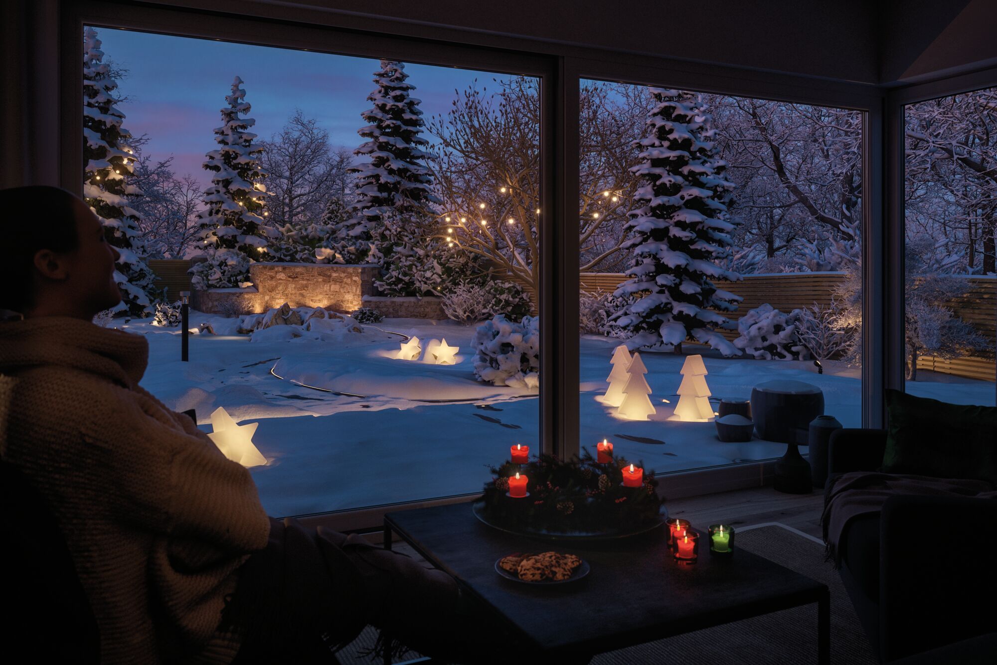 Wintergarten mit Kerzen und LED-Dekoleuchten in Stern- und Tannenform im verschneiten Garten bei Abenddämmerung