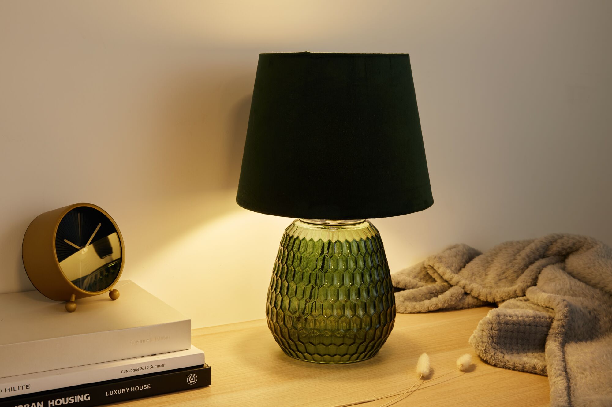 Table lamp with green glass base and black fabric shade on wooden surface near clock and blanket