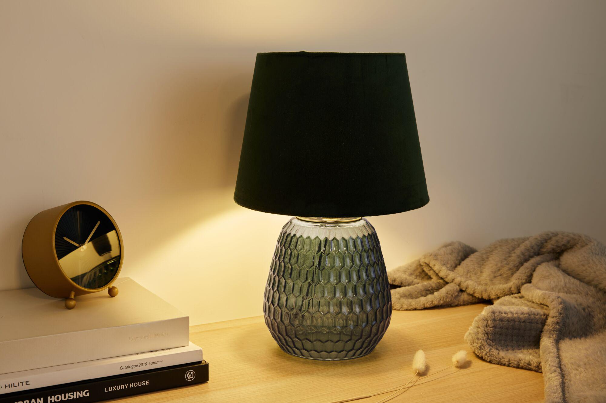 Tischlampe mit grünem Glasfuß und schwarzem Stoffschirm auf Holzoberfläche neben Uhr und Decke