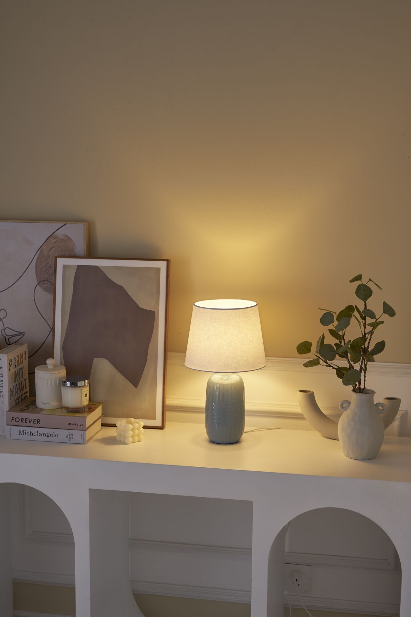 Lichtblauwe keramische tafellamp met witte stoffen kap op wit dressoir met decoratie en planten