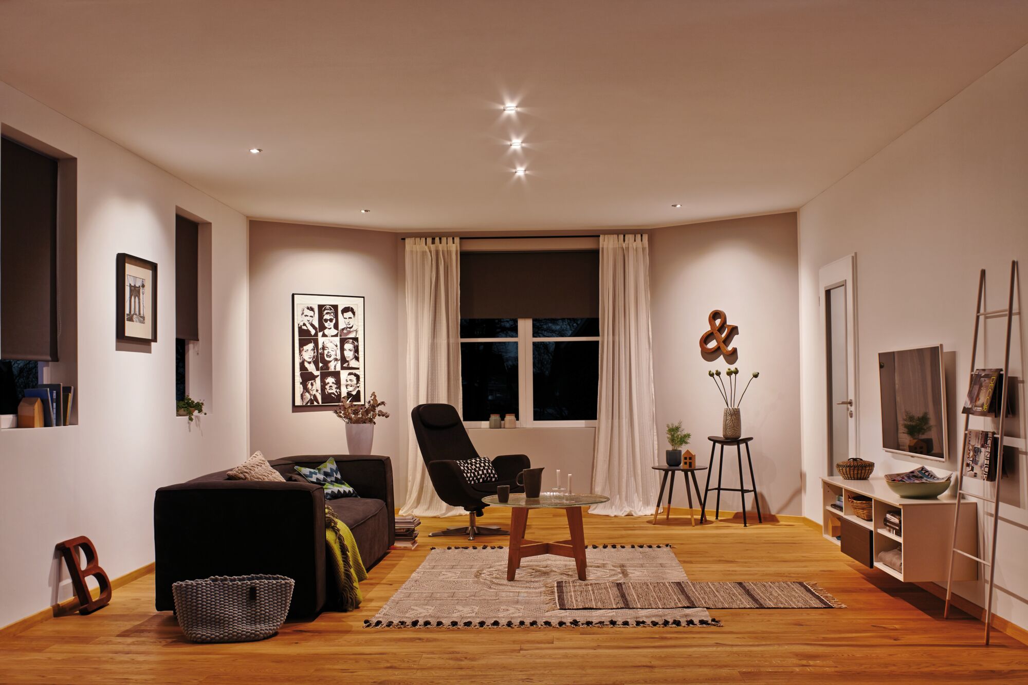 Modernes Wohnzimmer mit Holzfußboden, schwarzem Sofa, Sessel und warmweißer LED-Deckenbeleuchtung.