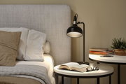 Black metal floor lamp beside bed with beige-grey headboard and bedside table with books and plant