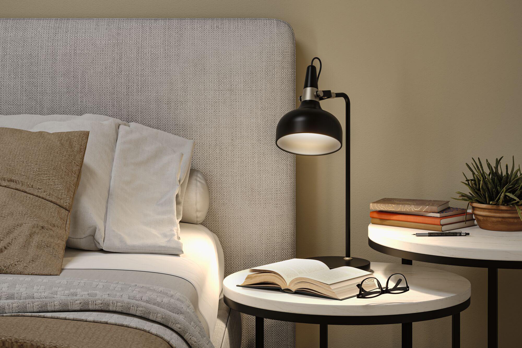 Black metal floor lamp beside bed with beige-grey headboard and bedside table with books and plant