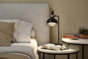 Black metal floor lamp beside bed with beige-grey headboard and bedside table with books and plant